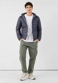 Veste matelassée gris foncé avec fermeture éclair devant, t-shirt blanc, pantalon cargo vert olive et baskets de couleur claire avec des accents.