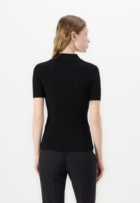 Haut noir à manches courtes à côtes avec un col polo, design ajusté et texture lisse, porté avec un pantalon noir sur un fond uni.