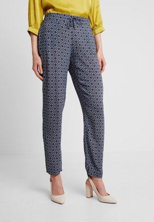 Mujer con pantalones ajustados azules con estampado, blusa amarilla de manga larga y zapatos de tacón beige, de pie sobre un suelo blanco.