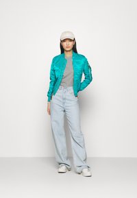 Teal satiinist bomber-jakk, hall t-särk, heledad sinised kõrge vöökohtadega teksad ja valged tennisetossud. Mudel kannab beeži mütsi. Lihtne taust.