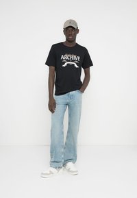 T-shirt preta com estampa "ARCHIVE" em branco, jeans azul claro e ténis brancos com detalhes em cinza, complementados por um boné bege.