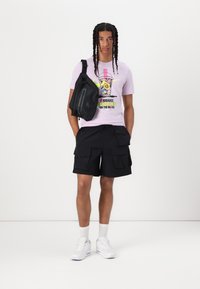 Black cargo shorts with side pockets, light purple graphic t-shirt featuring a drink design, and a black crossbody bag. White sneakers.