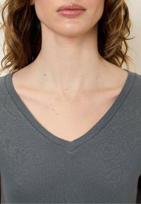 Šedý top s výstřihem do V, který má texturovanou tkaninu, přiléhavý střih a měkký límec. Tón pleti je viditelný kolem oblasti límce.
