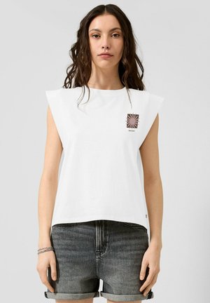 Femme portant un haut blanc sans manches avec un motif de soleil marron et le texte « Good Energy », associé à un short en jean noir retroussé.
