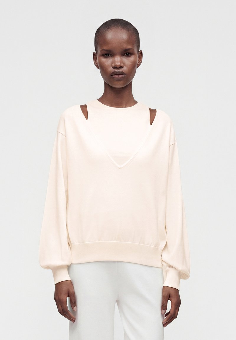 Personne aux cheveux courts portant un sweat crème avec des découpes triangulaires aux épaules et un pantalon blanc, debout devant un fond uni.