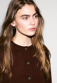 Tmavě hnědý pletený cardigan s kulatým výstřihem, se šedými knoflíky vpředu. Měkká textura a ležérní design.