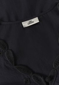 Mustast kangast pitsimist detailidega ääres ja valge riidetikandiga kaelaosas, millel on stiliseeritud logo.