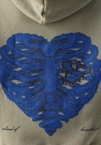 Felpa verde oliva con un grafico centrale di un design blu a forma di gabbia toracica con un cuore e accenti neri. Il testo recita "isola dei cuori spezzati".