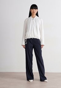 Woman stands against plain background wearing white zip-up blouse with gathered hem, wide black pants, and white pointed shoes.