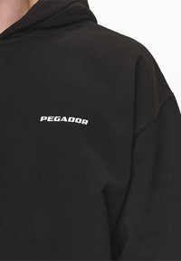 Felpa nera con cappuccio realizzata in tessuto morbido, con logo arcuato "PEGADOR" in bianco. Presenta un cappuccio con coulisse e polsini a costine.