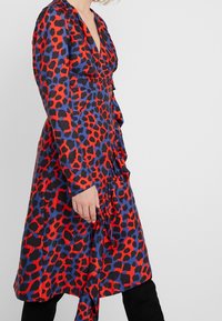 Långärmad klänning med ett rött och svart leopardmönster på en blå bakgrund, med en djup v-ringning och volangdetaljer fram.