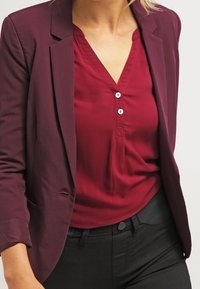 Blusa burdeos con cuello en v y tres botones blancos, usada debajo de una blazer a medida a juego. Presenta una textura suave y un diseño ajustado.