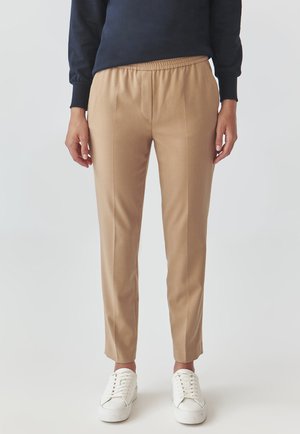 Beige, tailored pants with an elastic waistband, tapered leg design, and side seams. Paired with white sneakers.