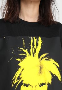 Černá mikina s výrazným žlutým motivem divokých vlasů na jednobarevném černém pozadí. Texturovaný žebrovaný výstřih.