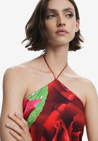Halter top med röd ros-mönster och gröna blad, med en knytkrage. Slät tyg med lätt lyster och vattendroppsdetailler.
