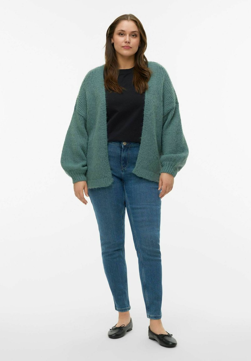 Cardigan vert surdimensionné et doux avec un tricot texturé, porté sur une chemise noire. Un jean skinny bleu et des ballerines noires complètent la tenue.