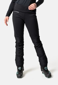 Pantalons isolés noirs au design ajusté, dotés d'une large ceinture et de genoux articulés. Associés à des boots de ski noirs avec des accents turquoise.