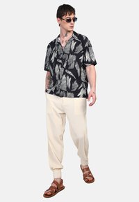 Camisa negra de manga corta con patrón de hojas blancas, pantalones a rayas crema y sandalias marrones con tiras. Lleva puesta una persona con gafas de sol y tatuajes.