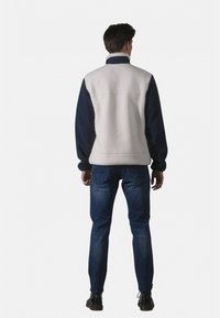 Veste en polaire blanche avec manches bleu marine, dotée d'un col montant et d'une coupe décontractée, associée à un jean foncé et des chaussures noires.