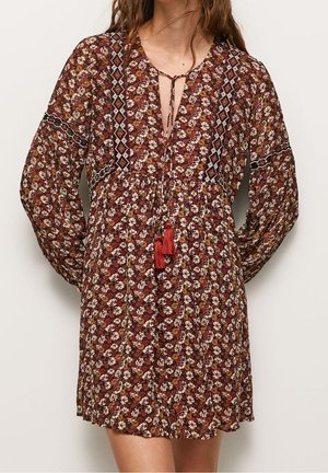 Mujer con un vestido marrón de manga larga con estampado floral, lazos de borlas rojas y ribete con patrón geométrico en las mangas y el frente.