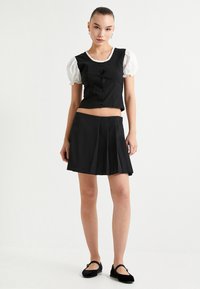 Schwarze, verkürzte Weste mit Knopfleiste, weißen Puffärmeln und passendem schwarzen Faltenrock. Weiche Textur, einfaches Design und minimalistischer Stil.