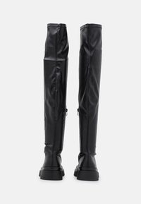 Bottes noires cuissardes en simili cuir lisse. Caractéristiques d'un design ajusté, semelles plates épaisses et fermetures éclair latérales discrètes.