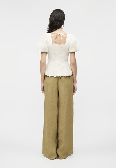 Γυναίκα με θέα από πίσω φοράει λευκό πουκάμισο με φουσκωτά μανίκια, δεμένο πίσω και με οδοντωτό τελείωμα, συνδυασμένο με φαρδιά παντελόνια από λινό σε ελαιώδες πράσινο χρώμα.