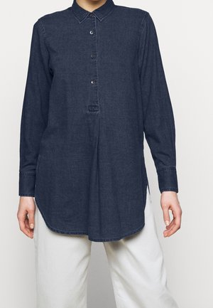 Kvinde iført en langærmet mørkeblå denimskjorte med knapper og hvide bukser, stående mod en ensfarvet lys baggrund.