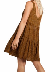 Mujer con un vestido marrón sin mangas, con un patrón de manchas negras, que presenta un espalda baja y una falda de capas, accessorios de anillos dorados.