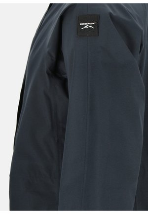 Veste imperméable bleu marine avec une texture lisse, arborant un patch noir avec "SWEDEMOUNT" sur le haut de la manche et un design épuré et minimal.