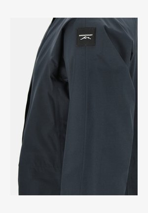 Veste imperméable bleu marine avec une texture lisse, arborant un patch noir avec "SWEDEMOUNT" sur le haut de la manche et un design épuré et minimal.