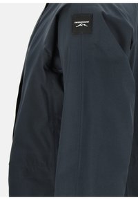 Marinblå vattentät jacka gjord av slät tyg med en märkeslogotyp i svart på överarmen och rena, minimalistiska linjer.