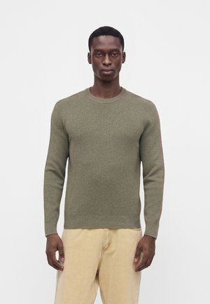Mand stående iført en olivengrøn langærmet sweater og beige bukser mod en ensfarvet hvid baggrund.