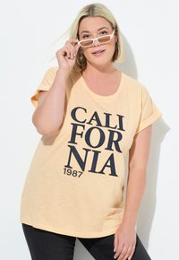 Světle broskvová tričko s ohrnutými krátkými rukávy, s výrazným námořnickým textem "CALIFORNIA 1987" v neformálním písmu. Hladká textura látky.