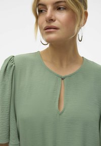 Šviesiai žalia bliuska su apvaliu iškirpimu, mygtuko kilpelės detalė ir minkšta, tekstūruota medžiaga; trumpi pūstieji rankovės suteikia apimties.