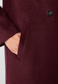 Mano nella tasca di un cappotto di lana rosso scuro con un bottone nero visibile nelle vicinanze, che mette in evidenza la trama del cappotto e i dettagli del design.