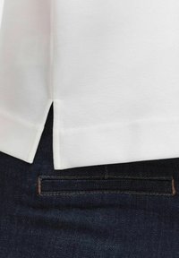 Valkoinen puuvillapaita suoremmalla helmalla ja sivu-aukoilla, yhdistetty tummansinihin farkkuihin, joissa on ommellut taskuyksityiskohdat.