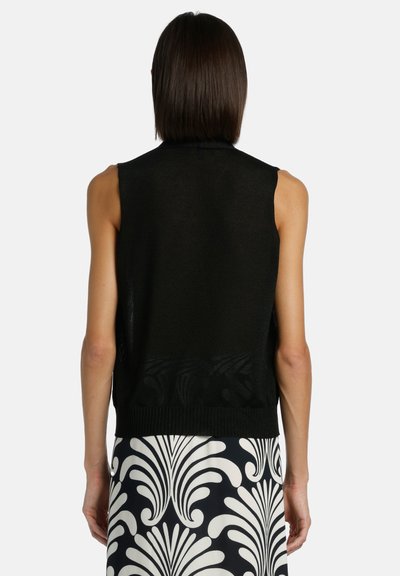 Femme aux cheveux bruns raides portant un haut noir sans manches et une jupe à motifs noir et blanc, vue de dos devant un fond uni.