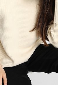 Cremfarbener, gerippter Pullover mit lockerer Passform und weiten Ärmeln, kombiniert mit schwarzen Hosen, die ein strukturiertes Strickmuster zeigen.