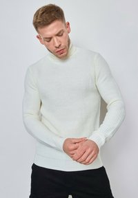 Pull en col roulé blanc, à côtes, avec des manches longues et une silhouette ajustée, fabriqué à partir d'un tissu tricoté texturé. Le modèle porte un pantalon noir.
