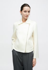 Hvid figursyet jakke med krave og guldfarvet lynlåsdetaile, med lange ærmer og blødt, tekstureret stof. Kombineret med grå bukser.