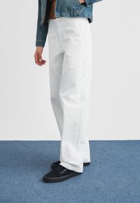 Άτομο που φορά λευκό φαρδύ παντελόνι, μαύρα αθλητικά παπούτσια και κοντό μπλε τζιν μπουφάν που στέκεται πάνω σε μπλε χαλί.