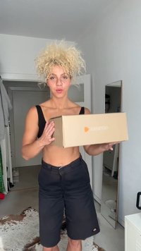 Brown cardboard box with "zalando" logo in orange, held in hands. Black shorts and sleeveless top worn by a person in a bright room.