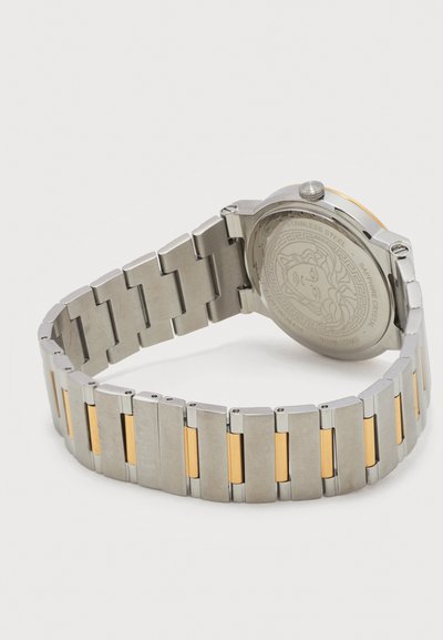 Nerezové hodinky so zlatými akcentmi na náramku, rytinou Medusy na zadnej strane a korunkou na boku.