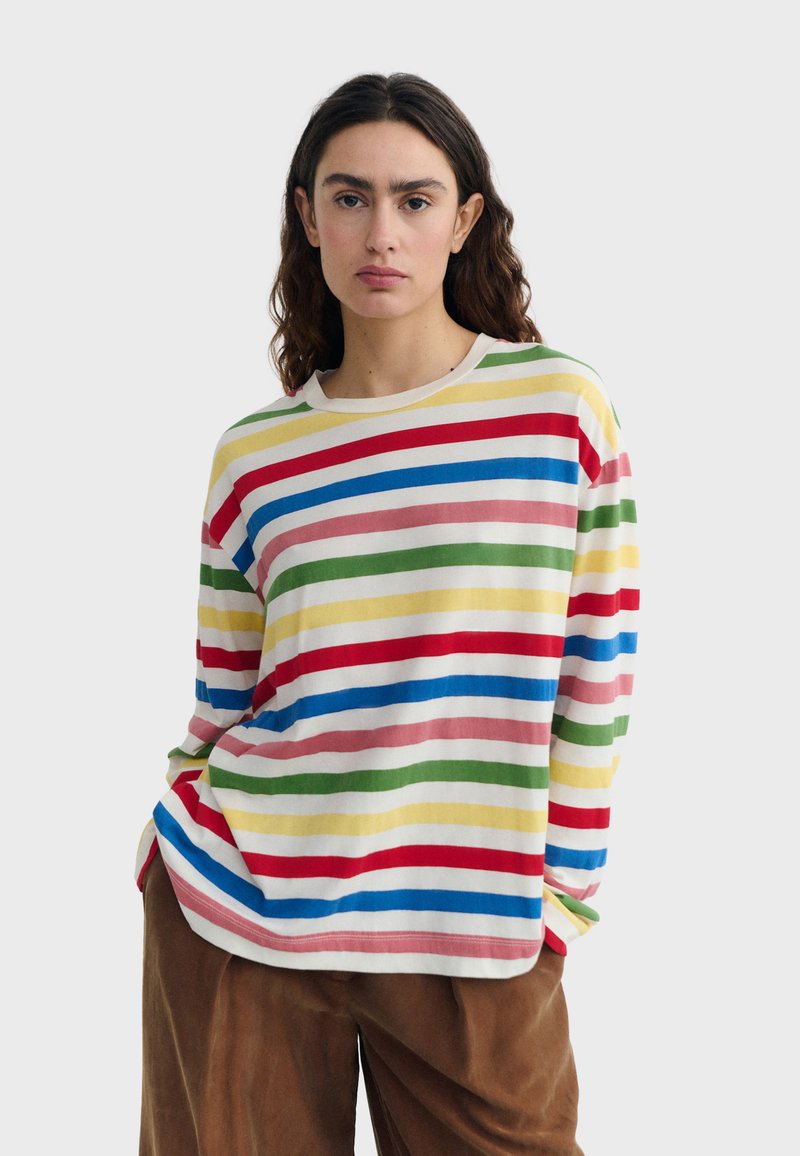 Femme aux cheveux ondulés portant une chemise à manches longues avec des rayures horizontales rouges, bleues, vertes, jaunes et roses, et un pantalon marron.
