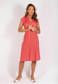 Rotes Baumwollkleid mit weißen Polka-Dots, kurzen Ärmeln und taillierter Brust. Der Rock schwingt leicht über dem Knie aus. Getragen mit weißen Absatzsandalen.