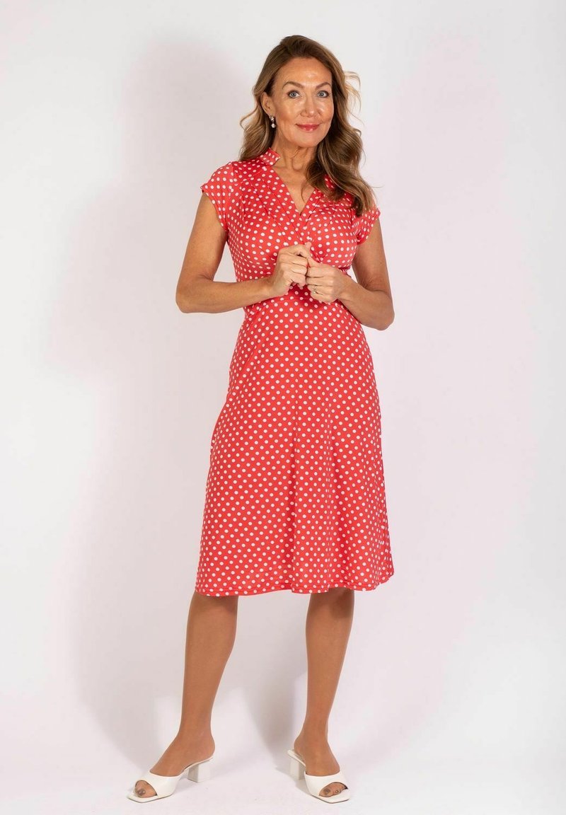 Rotes Baumwollkleid mit weißen Polka-Dots, kurzen Ärmeln und taillierter Brust. Der Rock schwingt leicht über dem Knie aus. Getragen mit weißen Absatzsandalen.