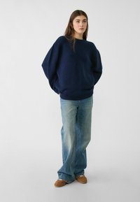 Temno modra prevelika pulover s okroglim izrezom, pletena tekstura in spuščena ramena, v kombinaciji s svetlo modrimi širokimi jeans hlačami in rjavimi čevlji.