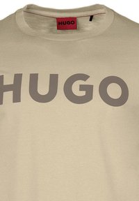 Béžové bavlněné tričko s kulatým výstřihem s velkým tmavě šedým nápisem "HUGO" přes přední část. Jednoduchá a hladká textura.