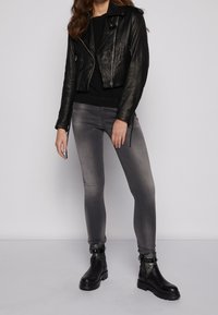 Chaqueta de cuero negra estilo biker, suéter negro, jeans ajustados grises y botas negras hasta el tobillo con detalle de hebilla, todo sobre un fondo liso.